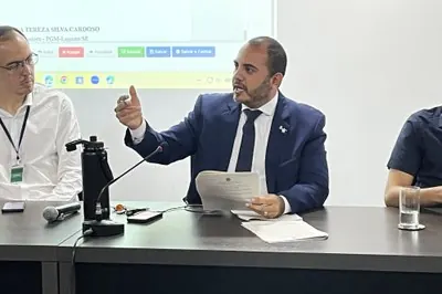 Fotografia em plano médio de um homem de terno azul-marinho, camisa branca e gravata escura, sentado à mesa durante uma reunião. Ele tem barba curta, segura papéis com a mão esquerda e gesticula com a mão direita erguida enquanto fala. Sobre a mesa à sua frente, estão dispostos um microfone de haste