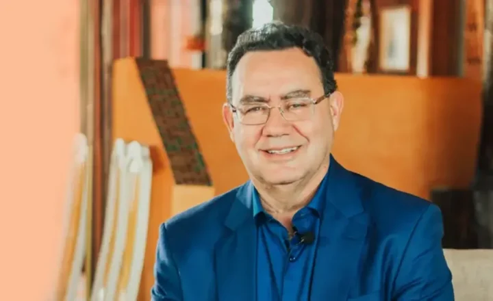 Um homem de meia-idade, Augusto Cury está sentado em um ambiente interno com iluminação quente, vestindo um terno azul sobre uma camisa azul mais escura e óculos de armação fina, sorrindo; ao fundo, vê-se uma luminária acesa e móveis de madeira.