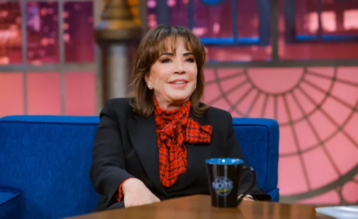 Mulher sentada em sofá azul em estúdio de programa de entrevista. Ela veste blazer preto e blusa com laço vermelho e preto, com caneca preta sobre mesa de madeira à frente.