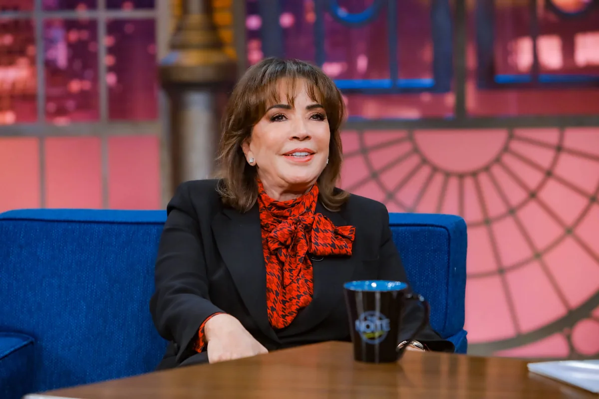 Mulher sentada em sofá azul em estúdio de programa de entrevista. Ela veste blazer preto e blusa com laço vermelho e preto, com caneca preta sobre mesa de madeira à frente.