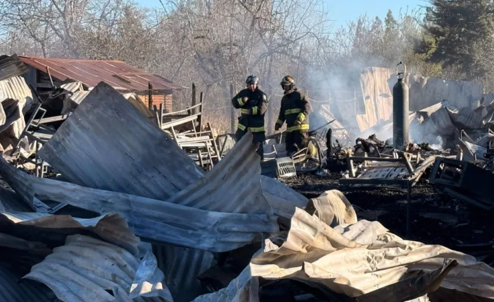 Dois bombeiros em uniforme completo estão em meio a destroços de estruturas metálicas retorcidas e queimadas, com fumaça ainda visível no ar. Ao fundo, árvores sem folhas e céu azul indicam ambiente rural em dia claro.