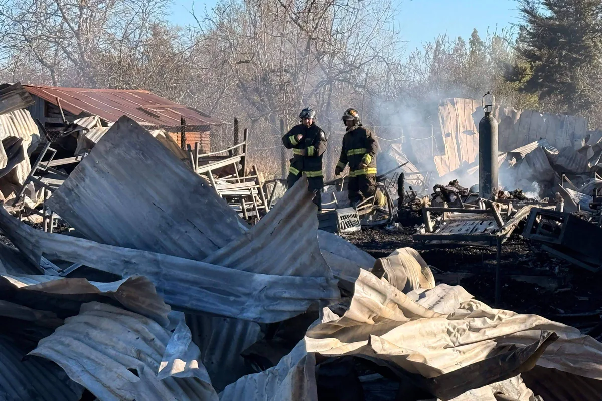 Dois bombeiros em uniforme completo estão em meio a destroços de estruturas metálicas retorcidas e queimadas, com fumaça ainda visível no ar. Ao fundo, árvores sem folhas e céu azul indicam ambiente rural em dia claro.