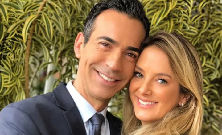 Um homem de terno azul-marinho e uma mulher de cabelos loiros sorriem, com os rostos próximos, em frente a uma parede de folhagens verdes e amarelas