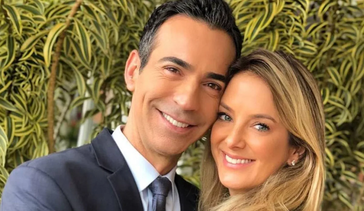 Um homem de terno azul-marinho e uma mulher de cabelos loiros sorriem, com os rostos próximos, em frente a uma parede de folhagens verdes e amarelas