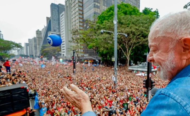 Lula, de perfil, sorri com os olhos fechados, vestindo camisa jeans azul, com a mão direita estendida para uma multidão de apoiadores em uma avenida urbana, com prédios altos e um balão azul ao fundo