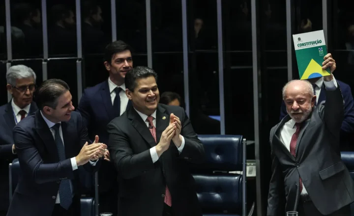 Homem de terno cinza levanta exemplar da Constituição brasileira em sessão parlamentar. Três homens ao redor aplaudem, todos vestidos formalmente, em ambiente institucional com cadeiras e grades ao fundo.