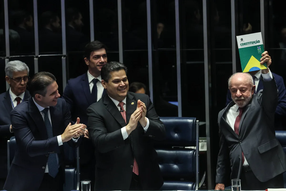 Homem de terno cinza levanta exemplar da Constituição brasileira em sessão parlamentar. Três homens ao redor aplaudem, todos vestidos formalmente, em ambiente institucional com cadeiras e grades ao fundo.