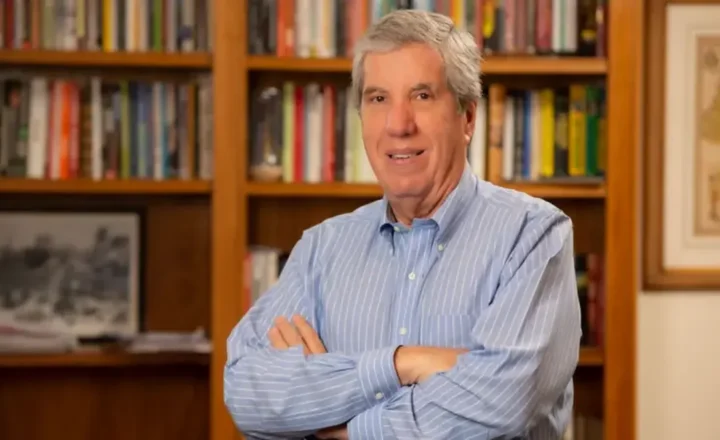 Fábio Coletti Barbosa, um homem de camisa social azul com listras brancas, de cabelos grisalhos, está de braços cruzados em frente a uma estante de livros