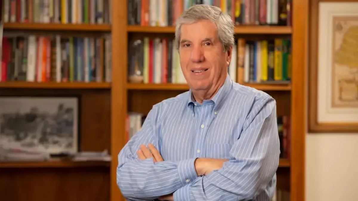 Fábio Coletti Barbosa, um homem de camisa social azul com listras brancas, de cabelos grisalhos, está de braços cruzados em frente a uma estante de livros