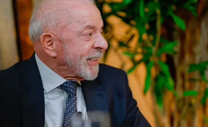 Homem branco idoso, de cabelos e barba grisalhos, sorrindo e olhando para a direita, vestindo terno azul-marinho, camisa social branca e gravata azul estampada, com plantas verdes desfocadas ao fundo