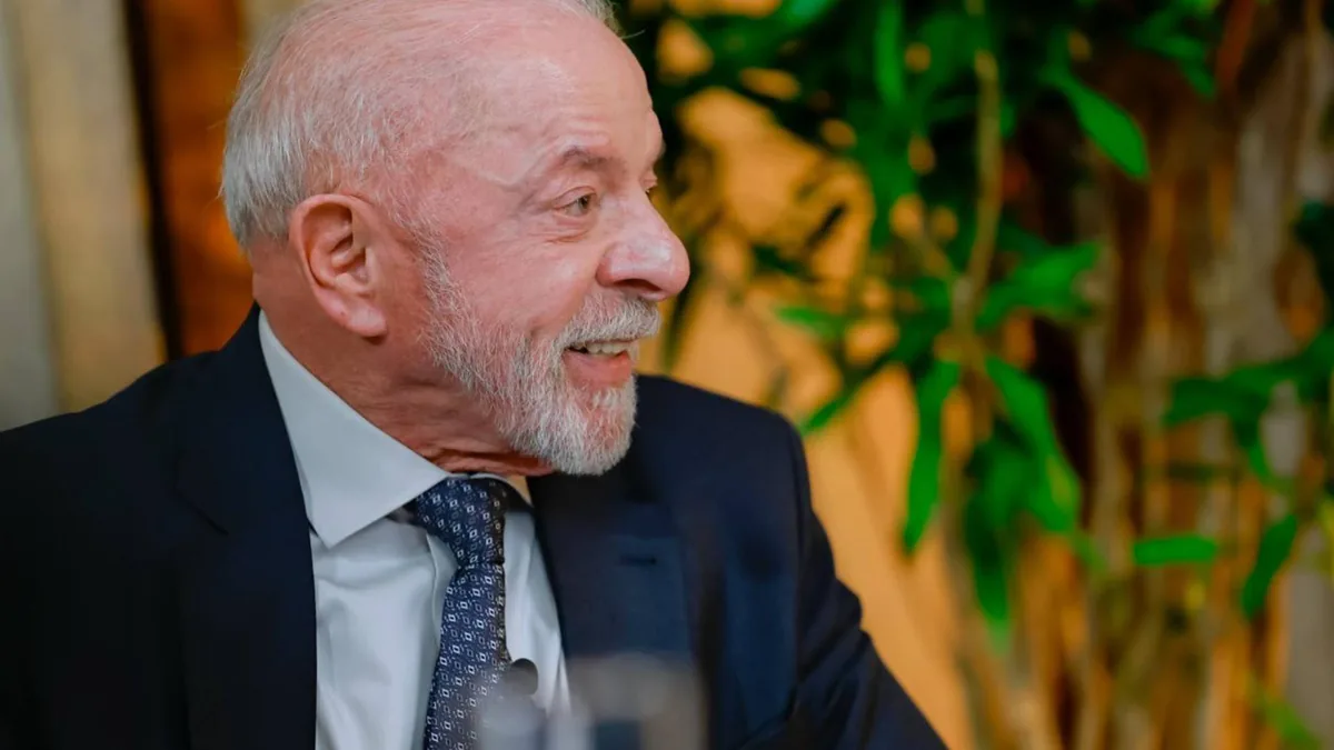 Homem branco idoso, de cabelos e barba grisalhos, sorrindo e olhando para a direita, vestindo terno azul-marinho, camisa social branca e gravata azul estampada, com plantas verdes desfocadas ao fundo