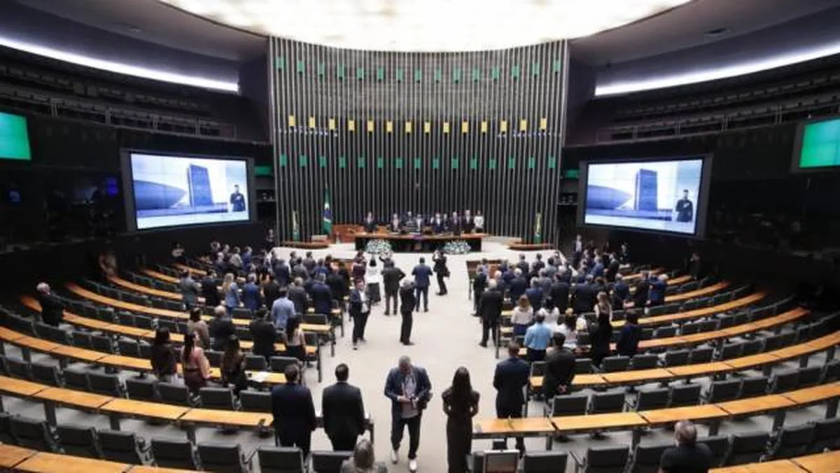 Plenário de um congresso com dezenas de pessoas em pé, algumas sentadas nas cadeiras de madeira e estofamento escuro. Ao fundo, uma mesa com autoridades e duas telas exibindo um homem e um prédio.