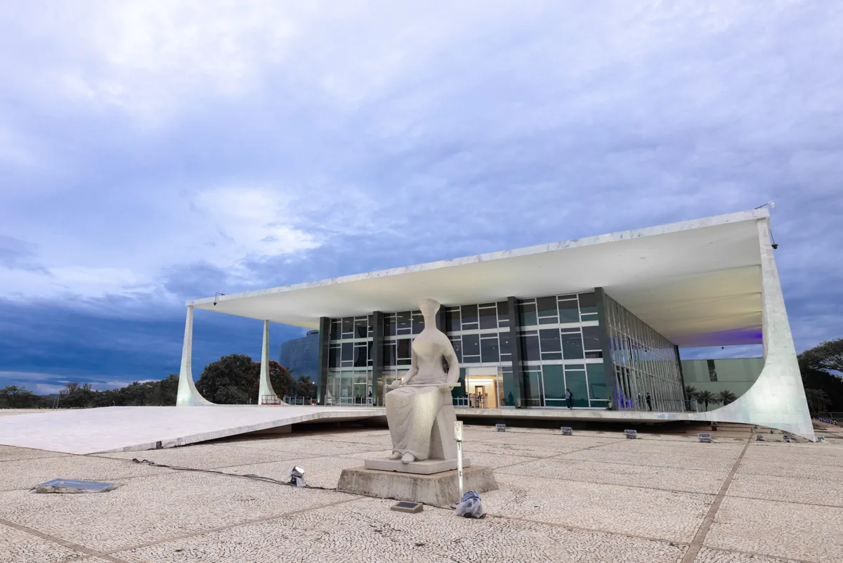 Foto em formato paisagem do prédio do STF ao entardecer e a Estátua da Justiça em primeiro plano