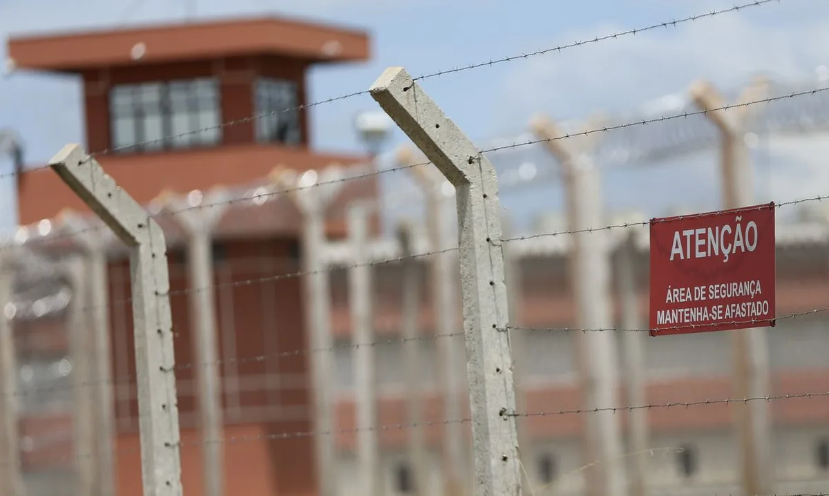 Fotografia de uma cerca de arame farpado de um presídio. À esquerda, uma placa com o texto