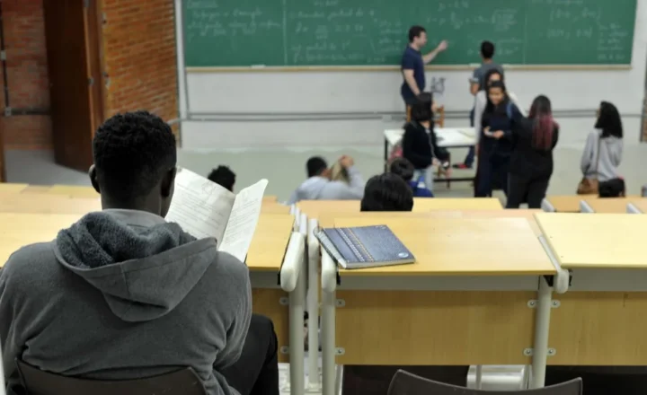 Fotografia de uma sala de uma universidade. Em primeiro plano, à esquerda, um aluno negro, de costas, sentado, lendo um material durante da aula