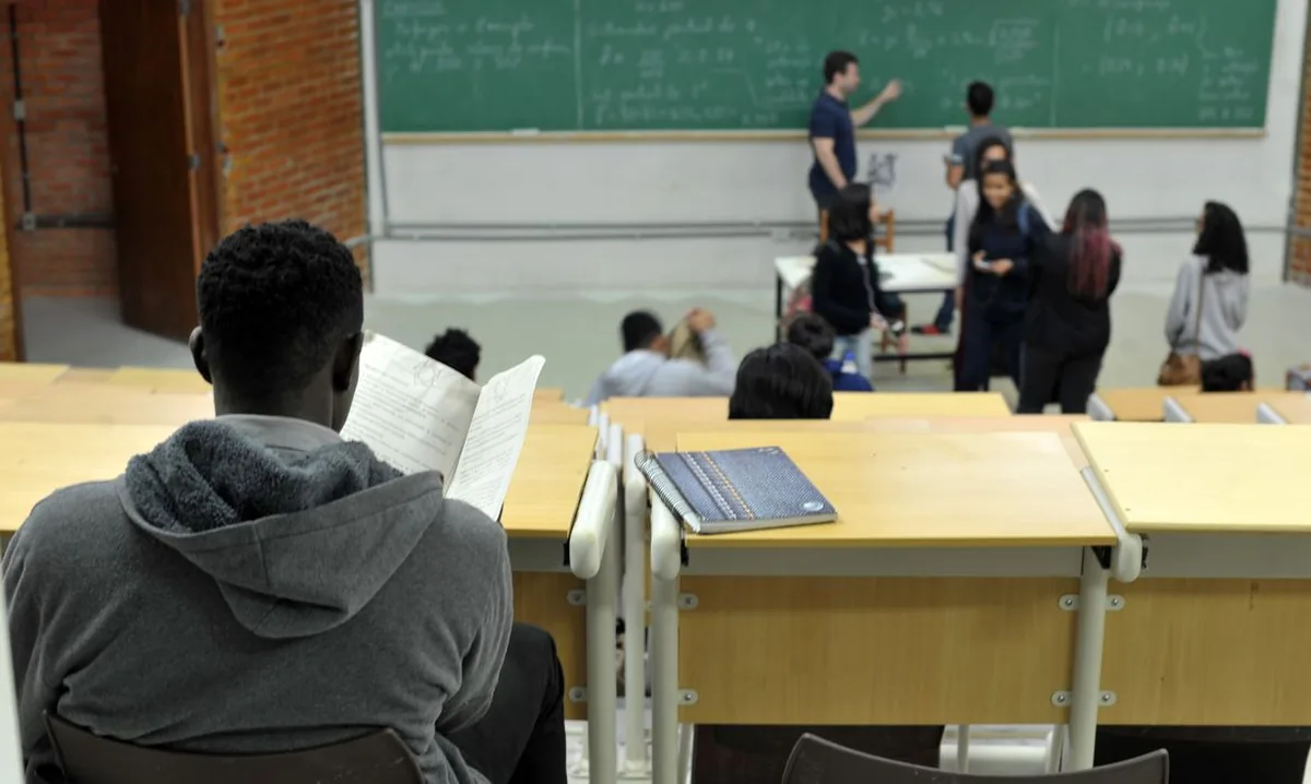 Fotografia de uma sala de uma universidade. Em primeiro plano, à esquerda, um aluno negro, de costas, sentado, lendo um material durante da aula