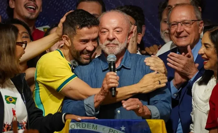 Grupo de pessoas reunidas em um palco ou evento público. Luiz Inácio Lula da Silva segura um microfone enquanto André Janones, vestindo uma camisa amarela com detalhes em verde, o abraça. Ao redor, várias pessoas aplaudem e se aproximam do centro da cena. Na parte inferior da imagem, há uma bandeira