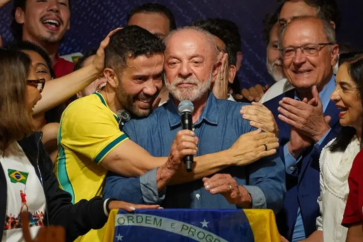 Grupo de pessoas reunidas em um palco ou evento público. Luiz Inácio Lula da Silva segura um microfone enquanto André Janones, vestindo uma camisa amarela com detalhes em verde, o abraça. Ao redor, várias pessoas aplaudem e se aproximam do centro da cena. Na parte inferior da imagem, há uma bandeira