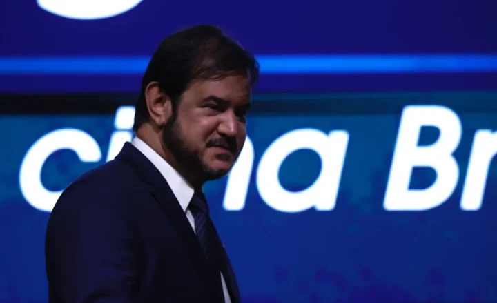 Homem de terno escuro e gravata está de perfil em ambiente escuro com fundo azul iluminado contendo texto parcialmente visível.