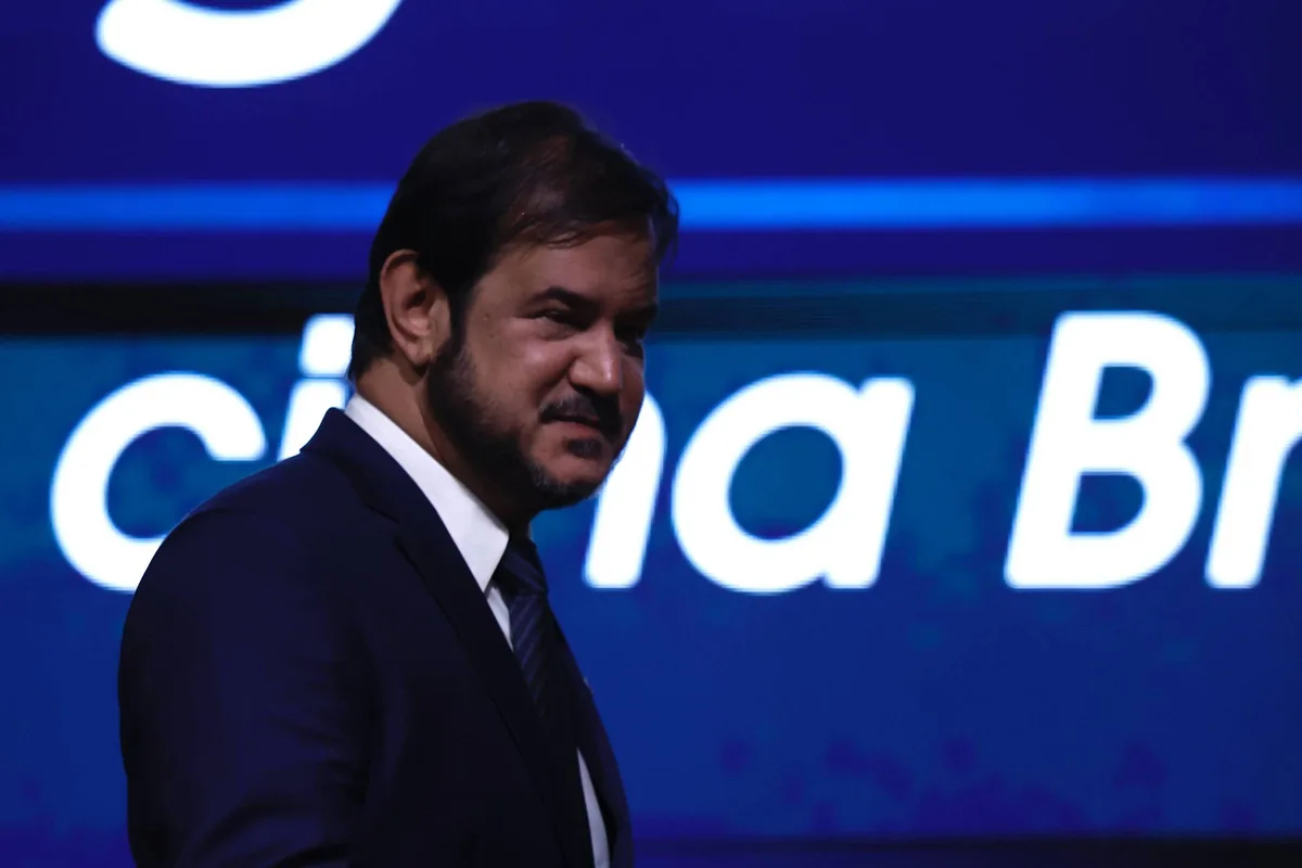 Homem de terno escuro e gravata está de perfil em ambiente escuro com fundo azul iluminado contendo texto parcialmente visível.